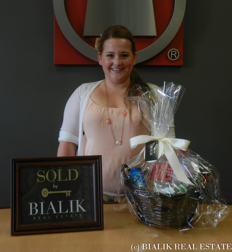 Smiling woman celebrates a home sale beside a SOLD sign and a wrapped gift basket.