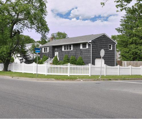 Dark shingle-sided corner-lot house behind a white picket fence and leafy trees.