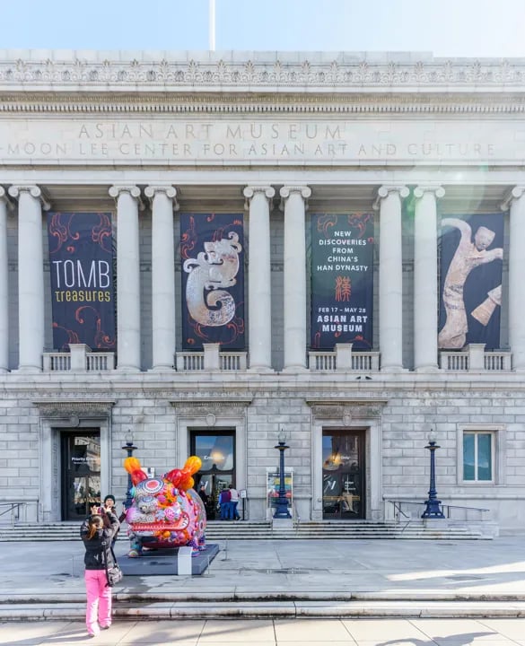 The grand entrance to the Asian Art Museum in San Francisco, featuring tall stone columns, exhibition banners, and a vibrant, multicolored sculpture.