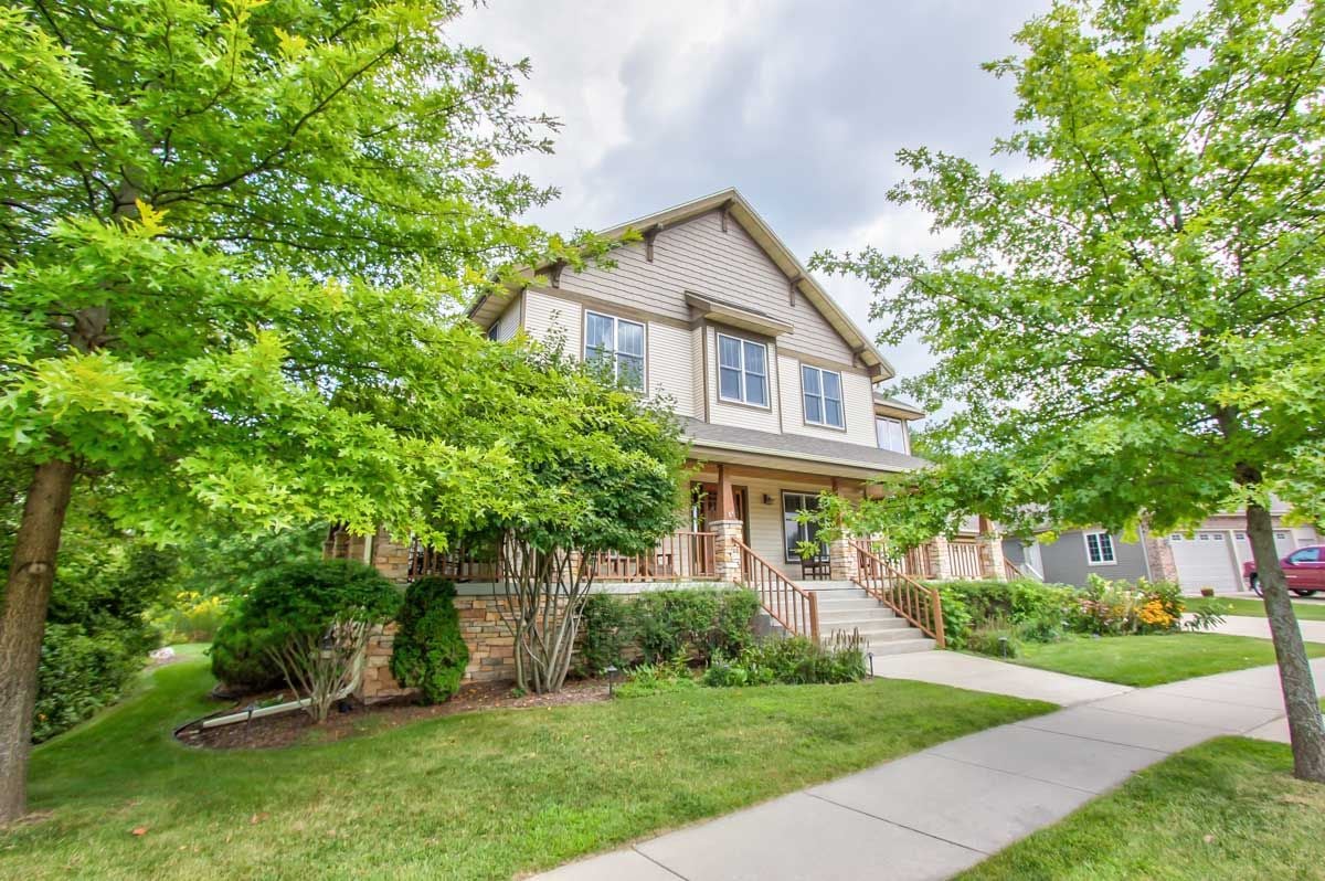 Two-story beige house with a covered porch, stone base, and leafy trees along the sidewalk.