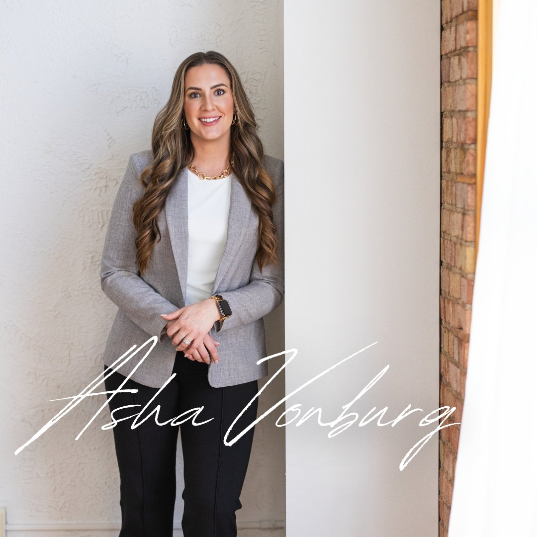 Asha Vonburg stands smiling in a gray blazer beside a textured wall and exposed brick.