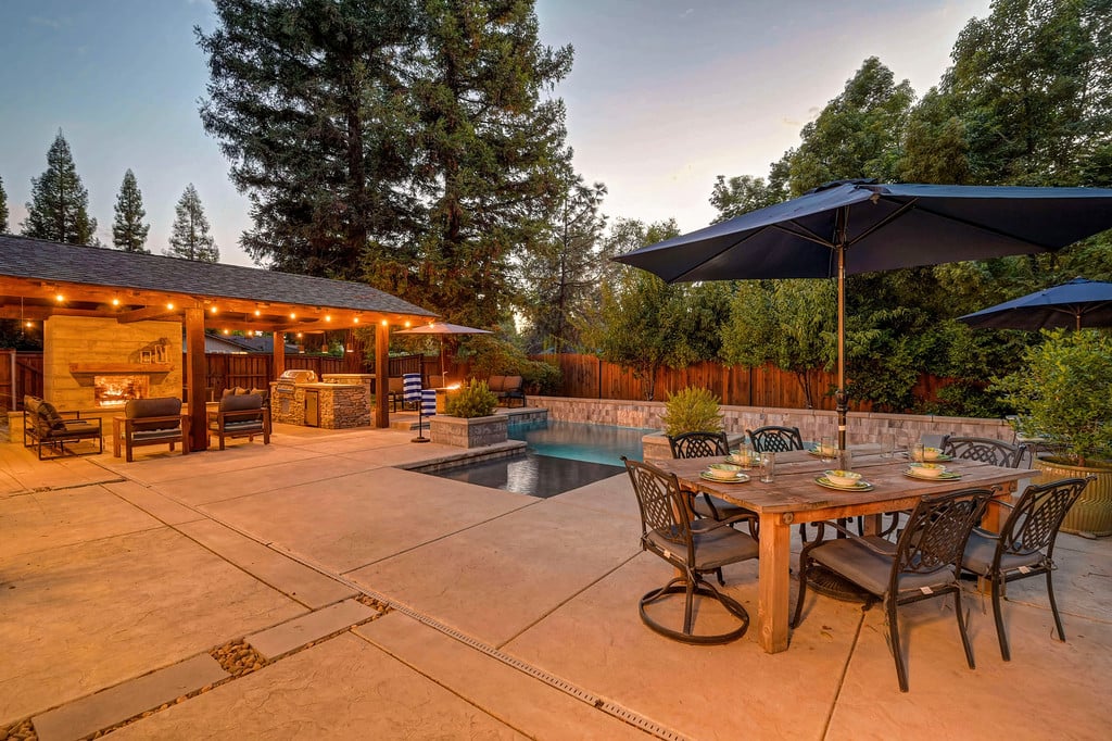 Backyard pool patio with covered fireplace seating and an umbrella-shaded dining table.