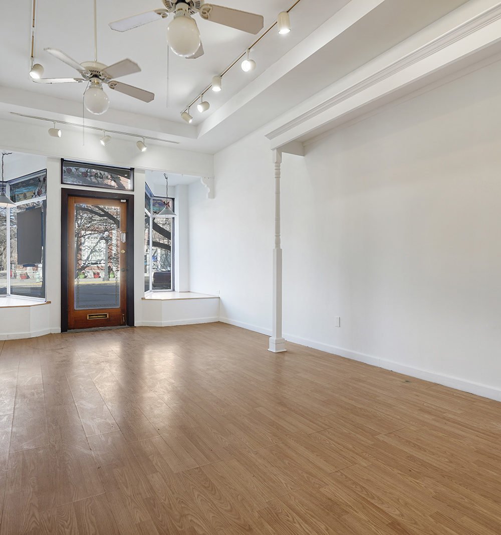 Vacant commercial interior with light wood floors, large front windows, and a glass entry door.