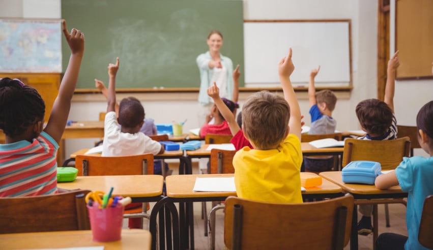 Children raise their hands at classroom desks as a teacher stands at the front.