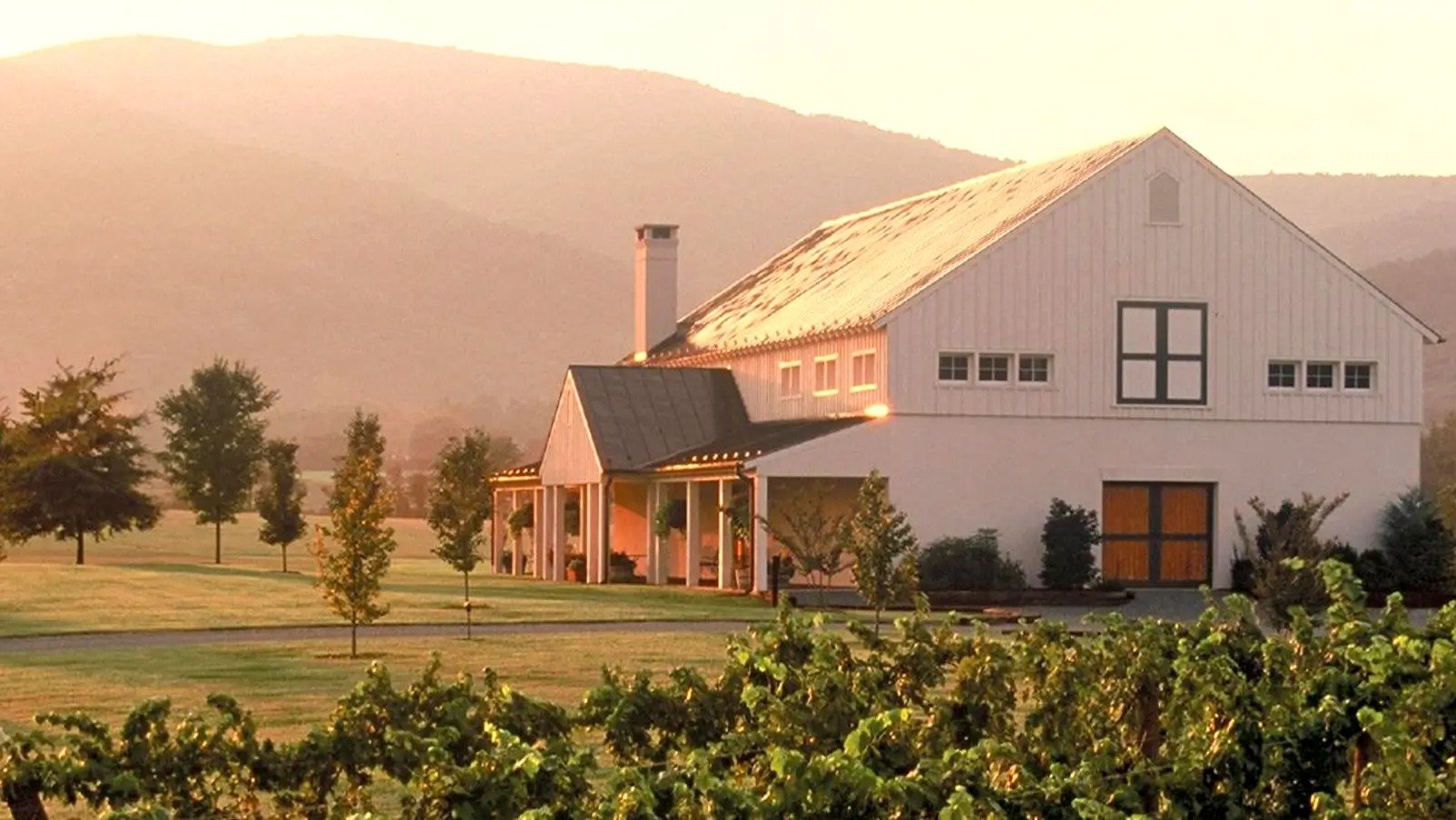 White barn-style building beside grapevines, with a mountain ridge in the background at sunset.