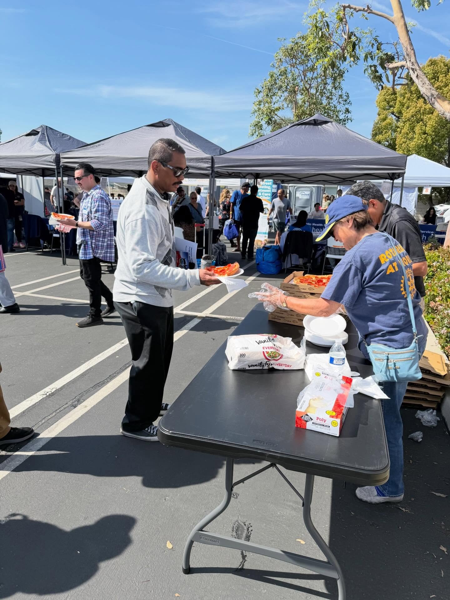 Volunteers serve pizza at an outdoor community fair beneath pop-up tents.