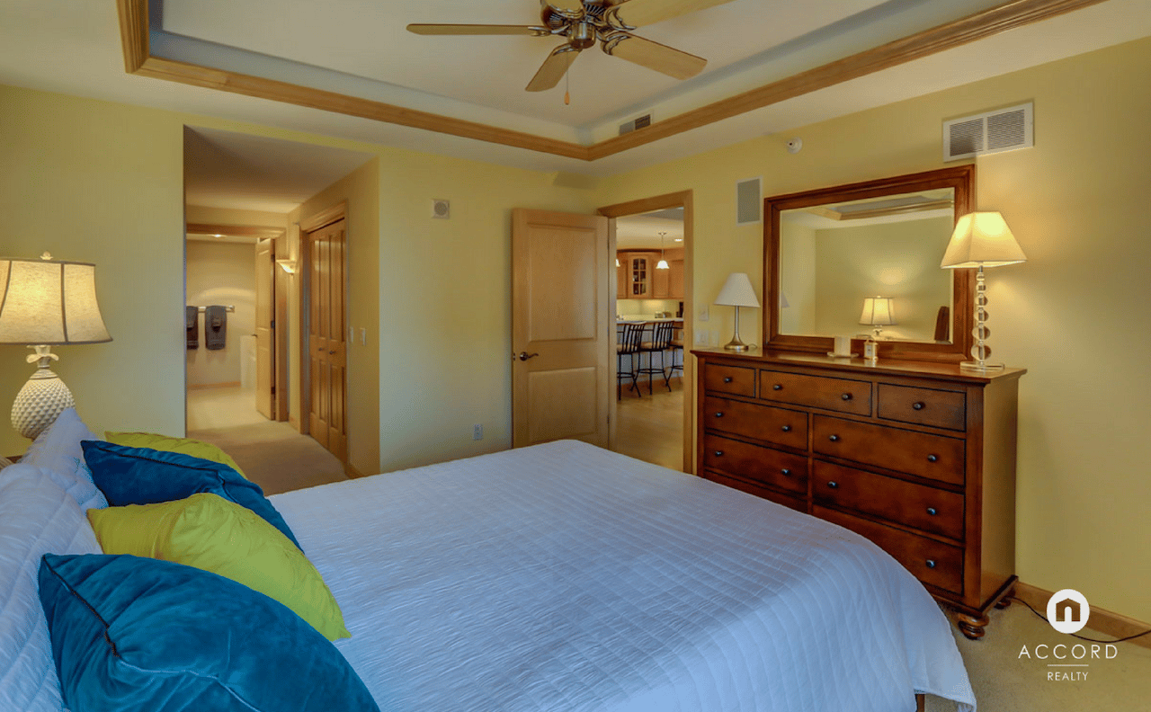 Furnished condo bedroom with a white bed, colorful pillows, and a wooden dresser beneath a large mirror.