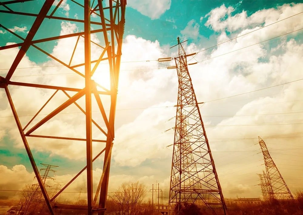 Electricity transmission towers and power lines silhouetted against a bright, cloudy sunset.
