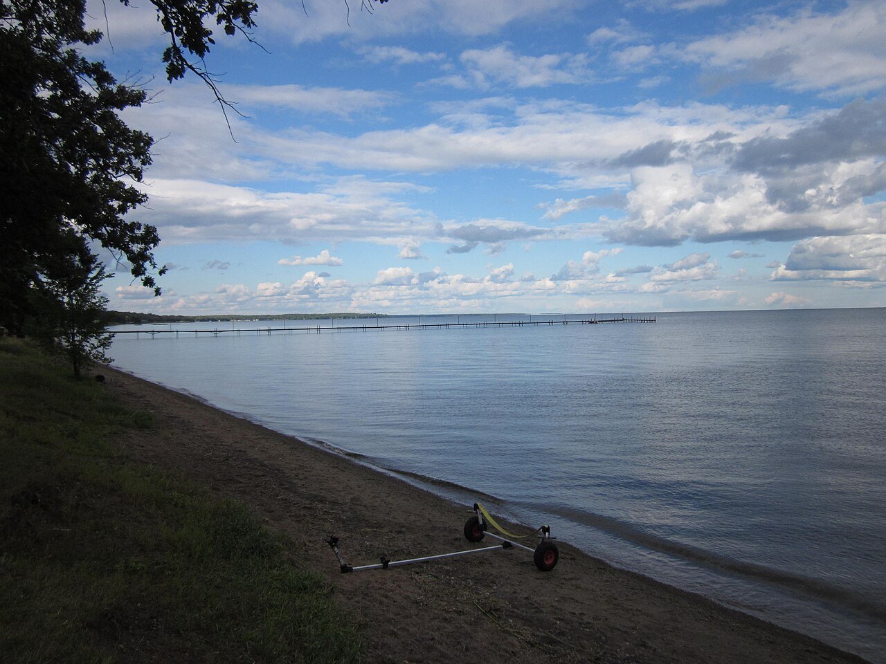 Mille Lacs Lake shoreline with a sandy beach, distant pier, and boat trailer.