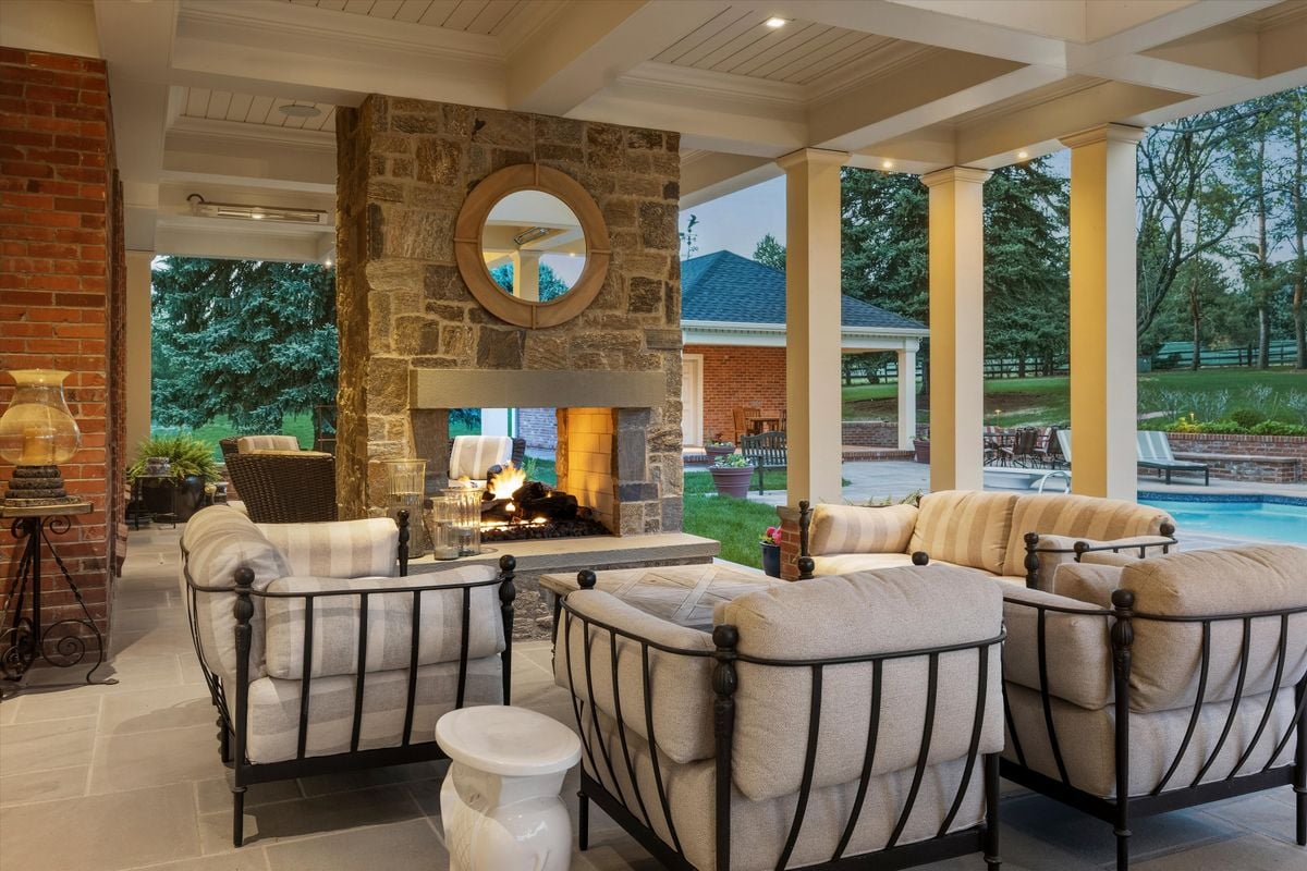 Covered patio with cushioned seating and a stone fireplace overlooking the yard and pool.