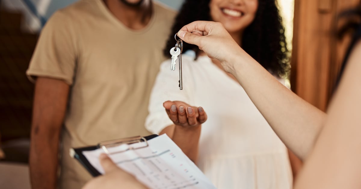 An over-the-shoulder view of a woman with a clipboard handing a set of keys to a couple at the front door of a house.