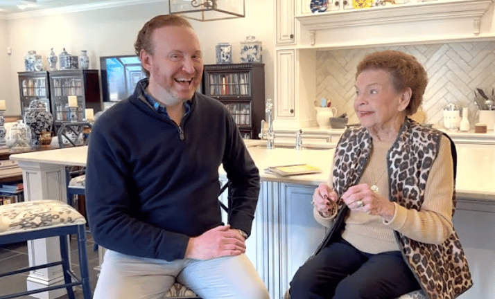A man and older woman chat while seated in a bright kitchen.
