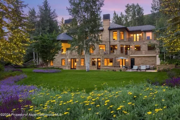 Stone-and-wood home glowing at dusk behind a lawn and flower garden.