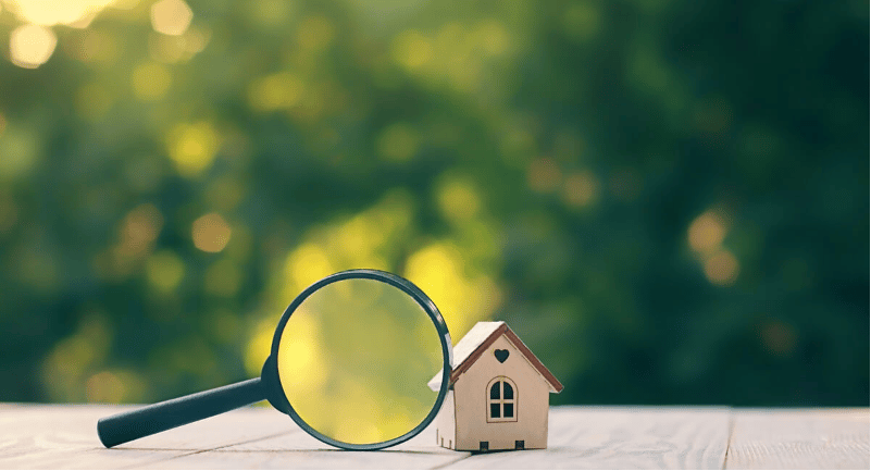 Miniature house beside a magnifying glass on a sunlit wooden surface.
