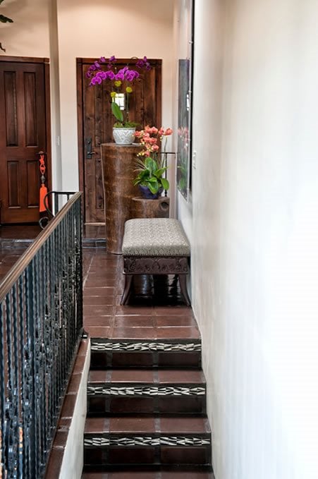 Hallway staircase with dark tiled steps, decorative metal railing, orchid-topped console, and upholstered bench.