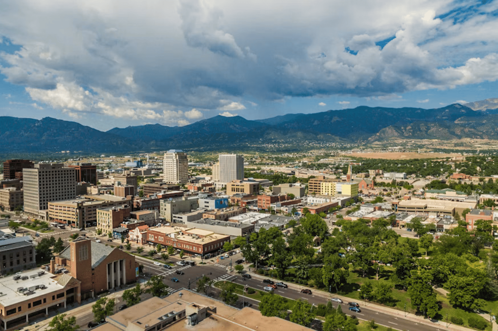 Downtown Colorado Springs and tree-lined streets with mountains beneath a cloudy sky.