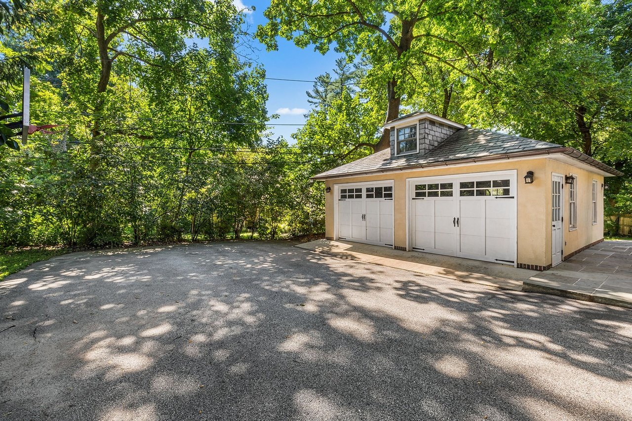 Detached beige garage with two white doors and a dormer, framed by mature trees beside a broad driveway.