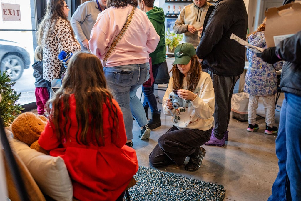 A woman in a Nest Realty cap kneels to photograph a child in a red dress at a gathering.