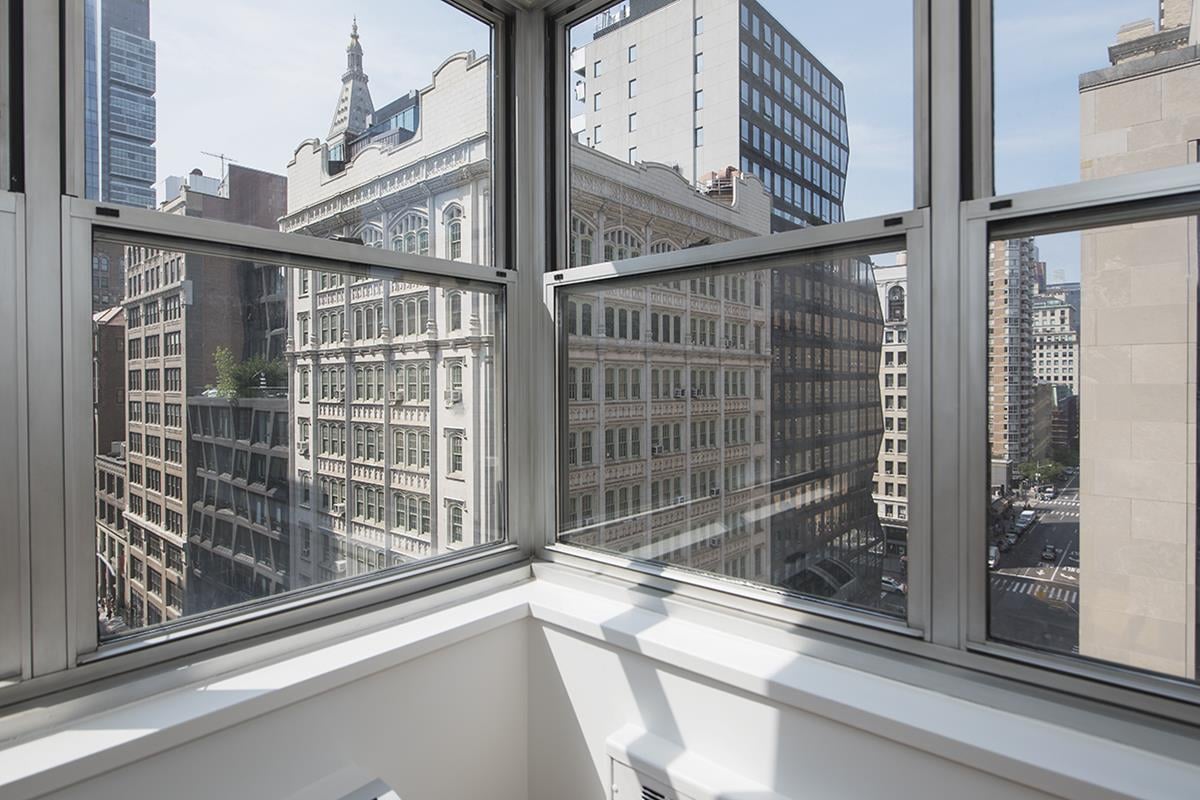 Wraparound apartment windows overlook Manhattan buildings, with sunlight falling across a white windowsill.