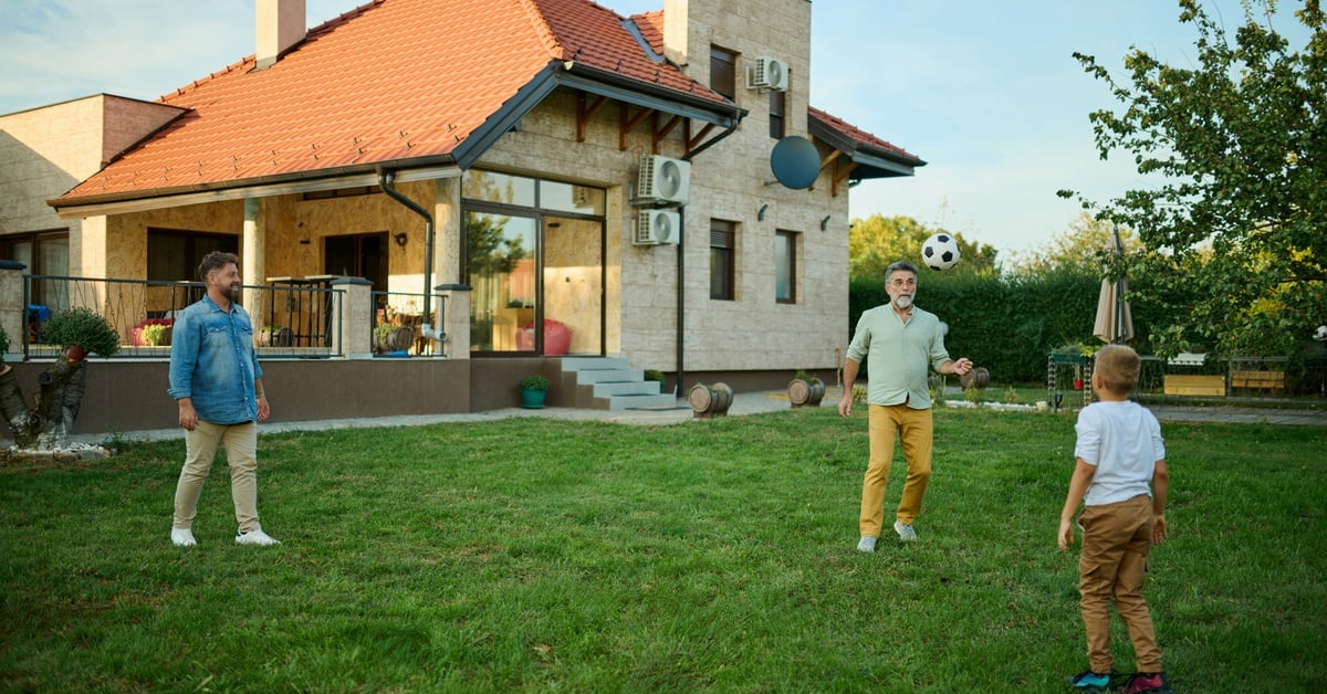 A man, an older man, and a young boy kicking a soccer ball between each other in the green backyard of a home.