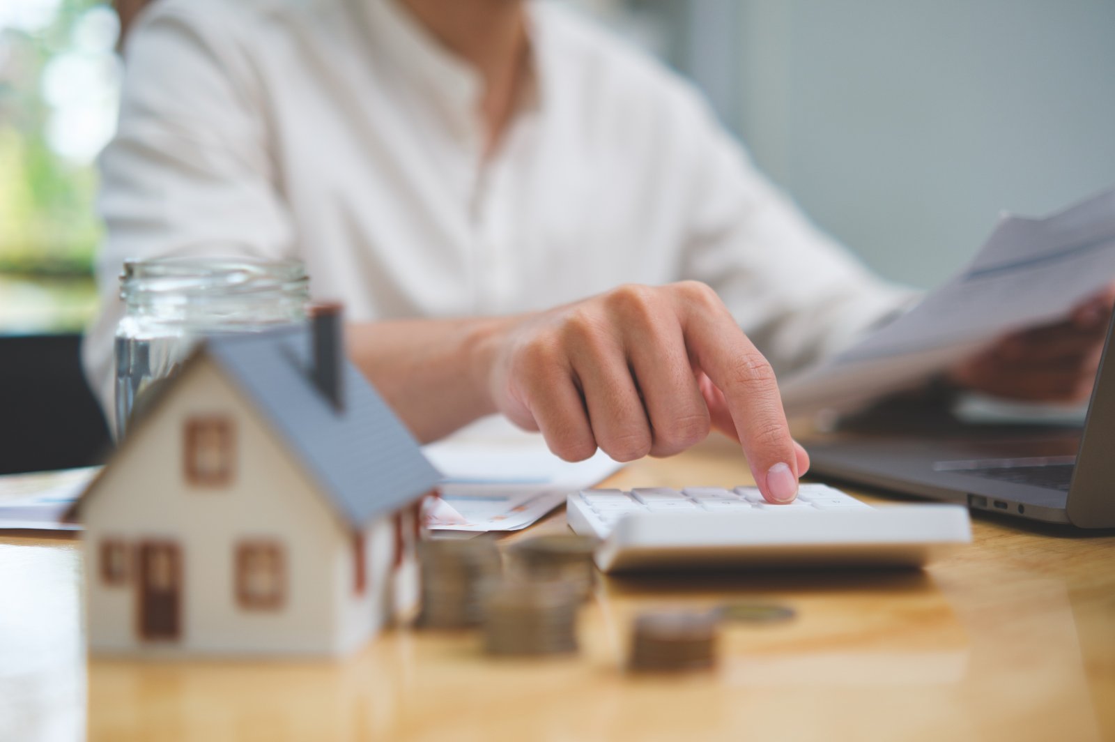 A person calculates real estate finances beside a model house, laptop, paperwork, and coins.