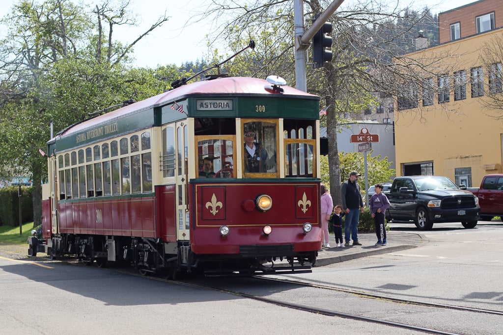 Historic red and cream streetcar traveling on tracks through downtown Astoria, Oregon, with pedestrians and Astoria buildings in the background
