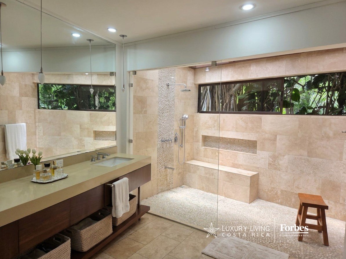 Luxury bathroom with a dark-wood vanity, glass shower, stone tile, and windows overlooking greenery.