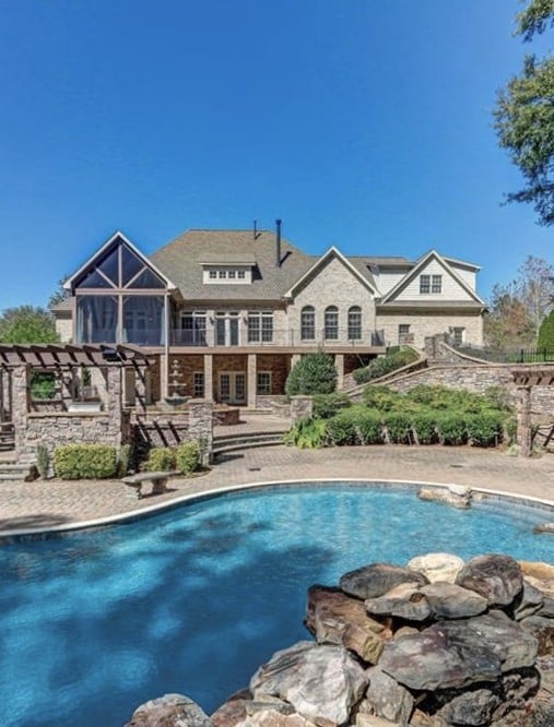 Large stone-and-brick home behind a curved pool, with a pergola and landscaped stone patio.