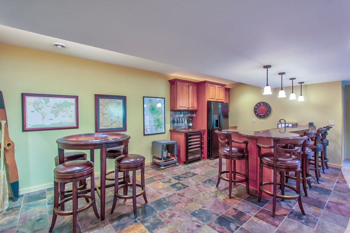 Home bar with reddish-wood cabinetry, leather stools, and colorful stone tile flooring.