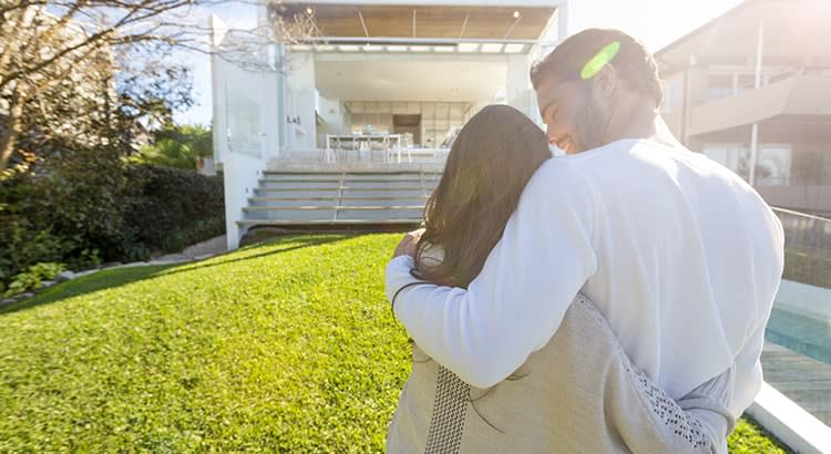  This image likely represents homeownership, purchasing a new home, or family life, as the couple shares a tender moment outdoors with a house in the background.