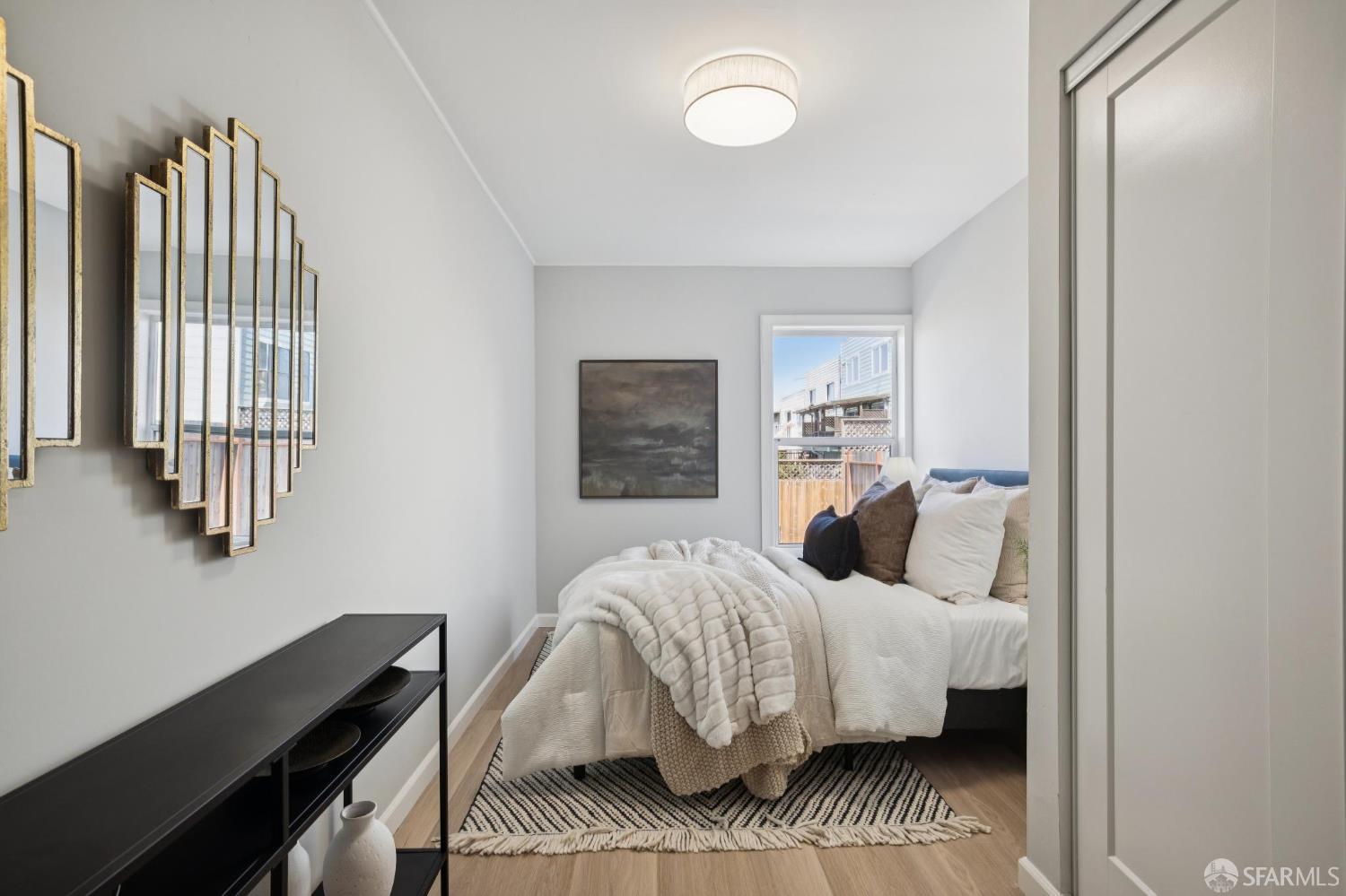 Staged bedroom with white bedding, geometric wall mirrors, and a window overlooking nearby homes.