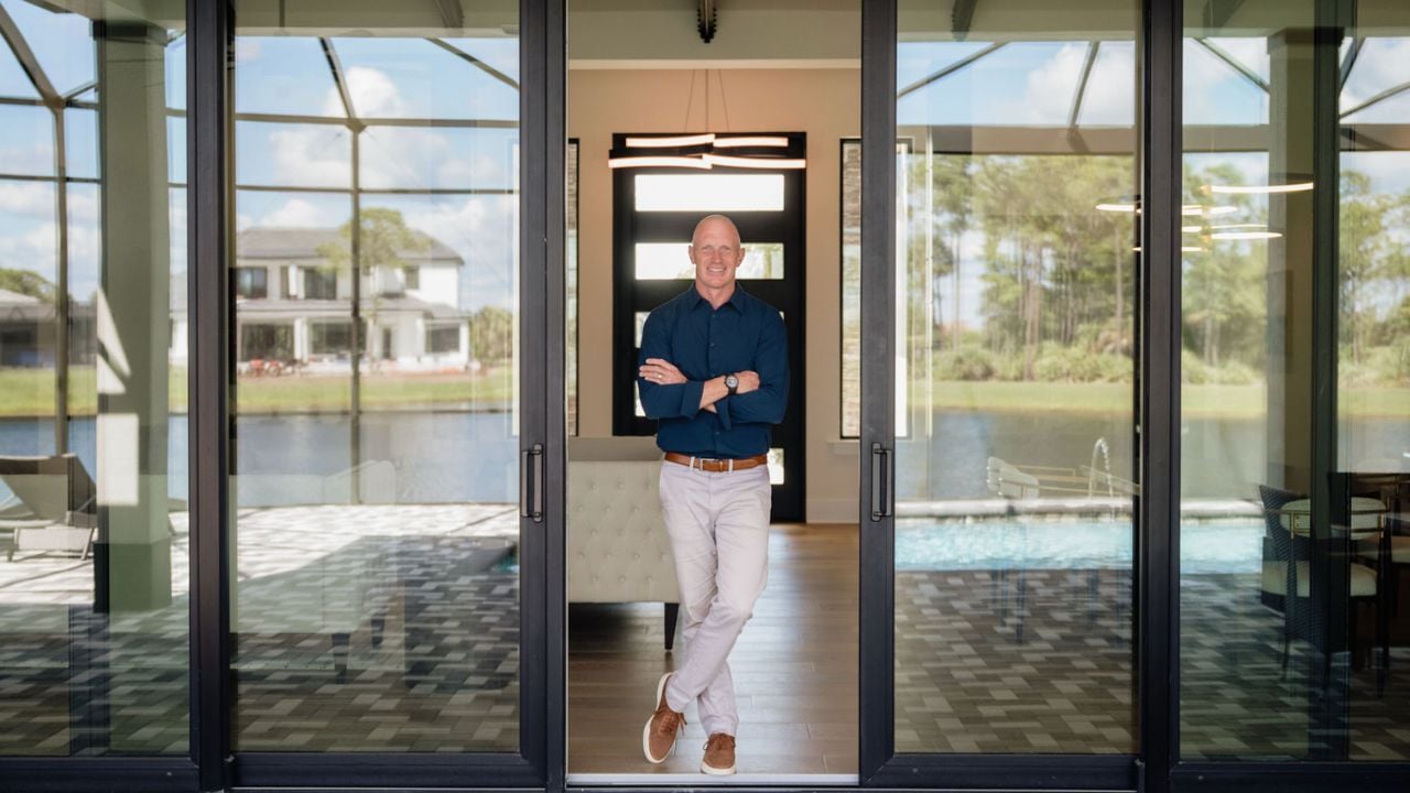 Man in a navy shirt stands in a glass doorway overlooking a pool.