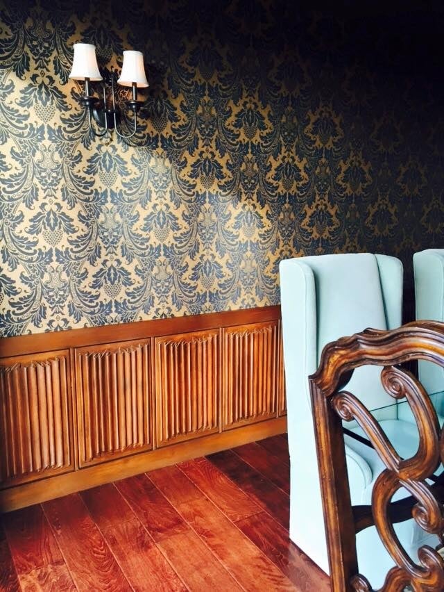 Vintage interior with a pale blue chair, damask wallpaper, carved wood paneling, and hardwood floors.