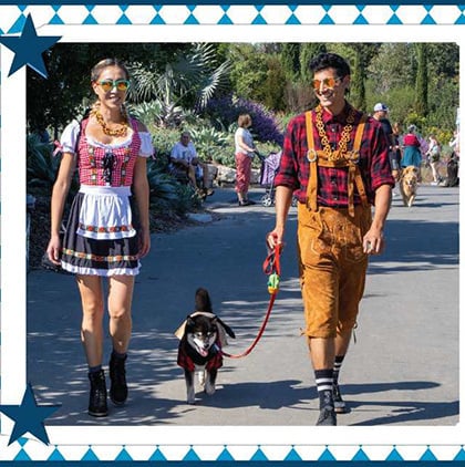 Two people in Oktoberfest outfits walk a dog along a garden path.