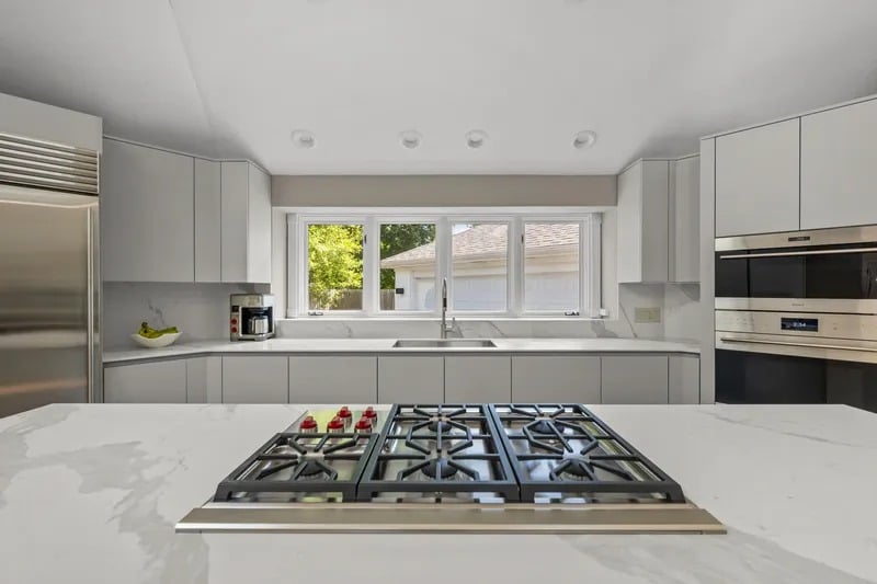 Modern white kitchen with a marble island, six-burner cooktop, and wide window above the sink.