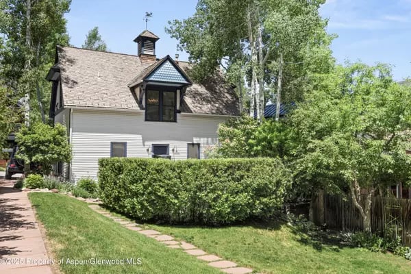 White-sided house with a steep shingle roof and dormer, partly screened by leafy trees and hedges.