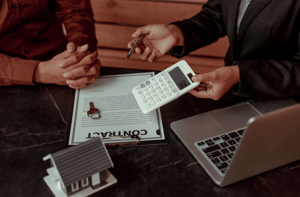 People review a real estate contract with a calculator, keys, laptop, and model house.