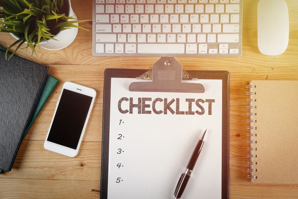A numbered checklist on a clipboard beside a keyboard, phone, notebooks, pen, and potted plant.