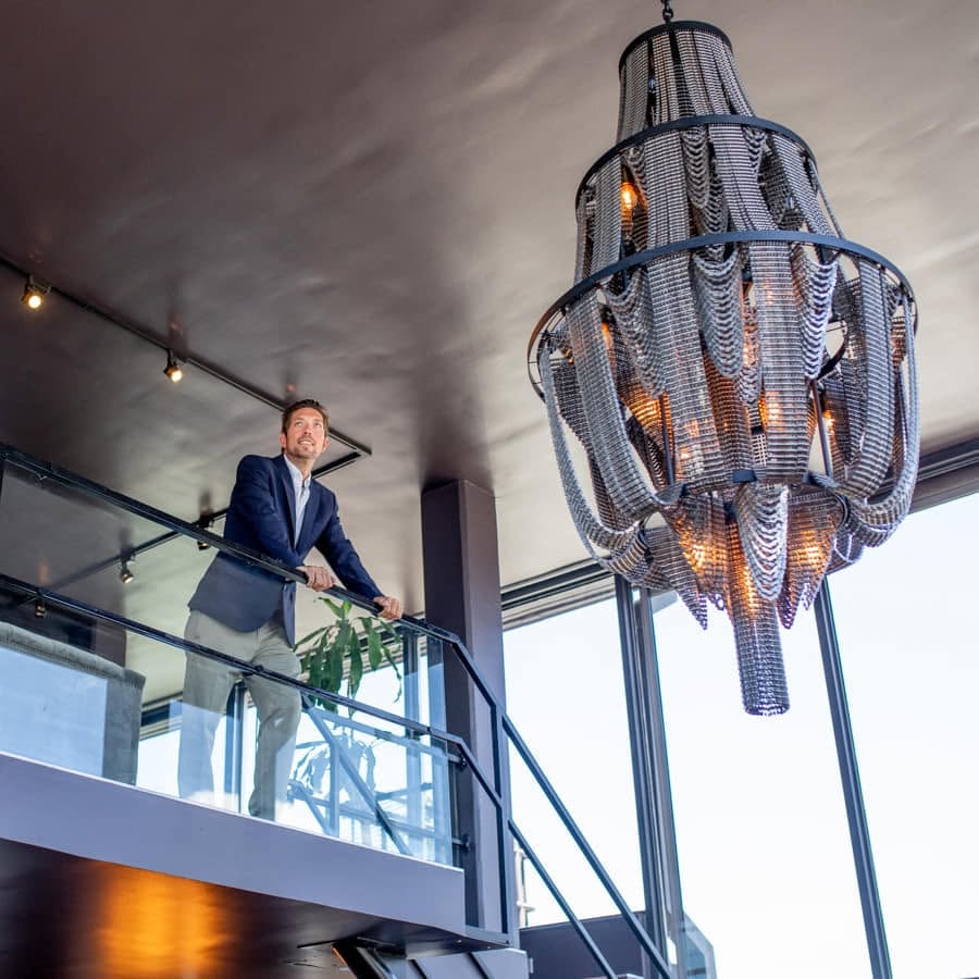Modern interior with a glass-railed mezzanine and oversized chandelier, with a man standing above.