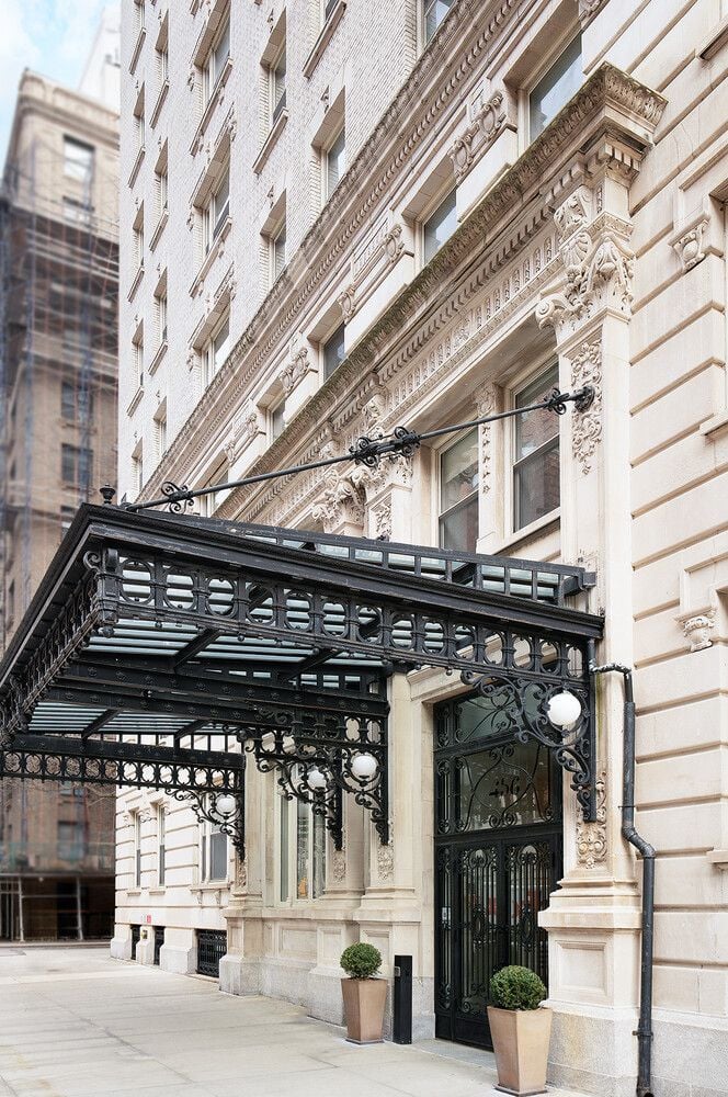 Apartment building entrance with an ornate stone facade, wrought-iron canopy, and potted shrubs.