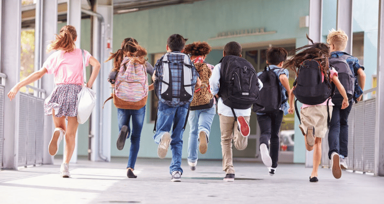 Schoolchildren with backpacks walk toward a building entrance along a covered walkway.
