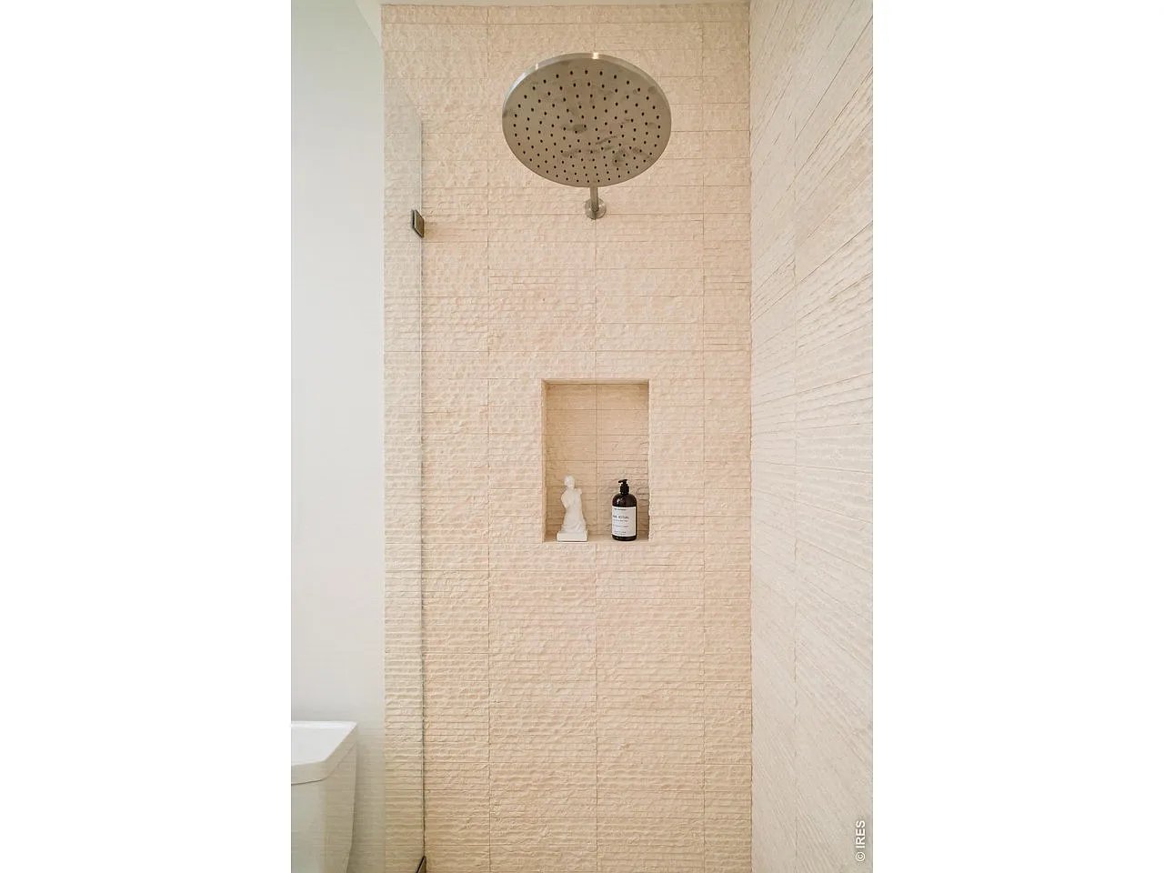 Walk-in shower with pale textured tile, a round rainfall showerhead, and a recessed shelf.