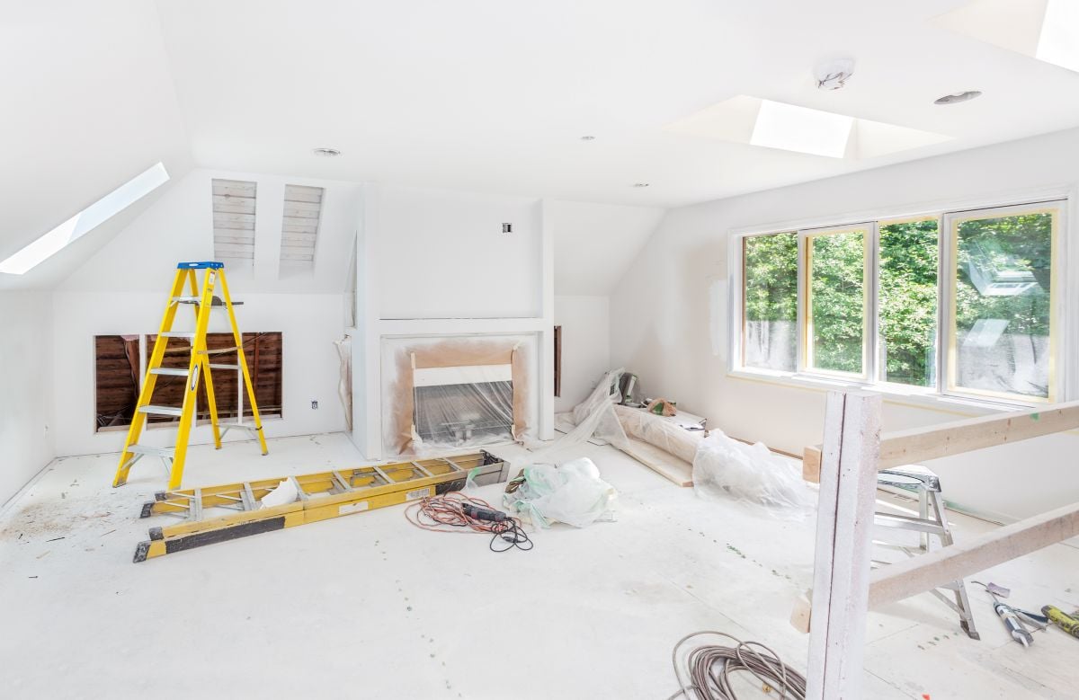 Attic living room under renovation with a fireplace, yellow ladders, tools, and large windows overlooking trees.