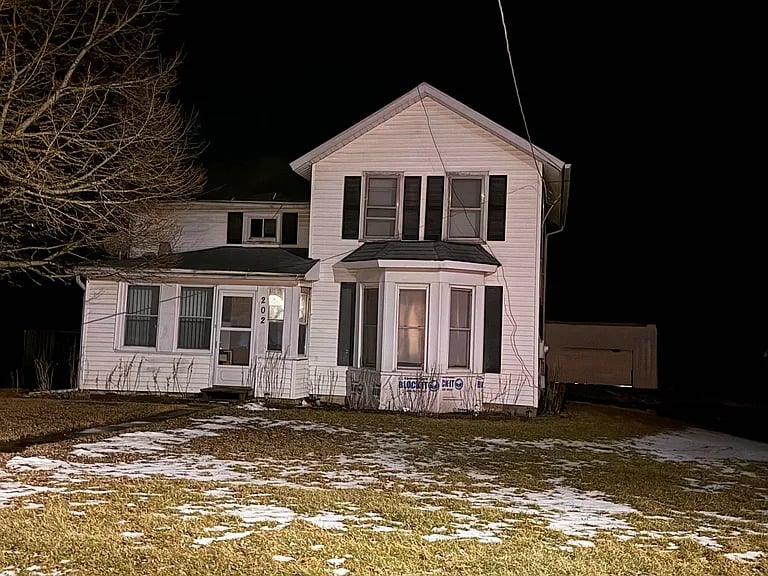 White two-story house with black shutters and a bay window, photographed at night with patchy snow in the yard.