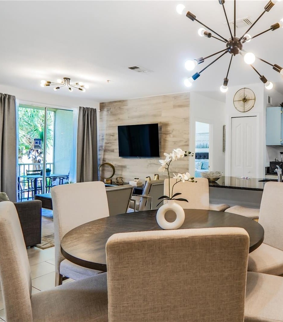 Open-plan dining and living area with a round table, branching chandeliers, and patio doors.