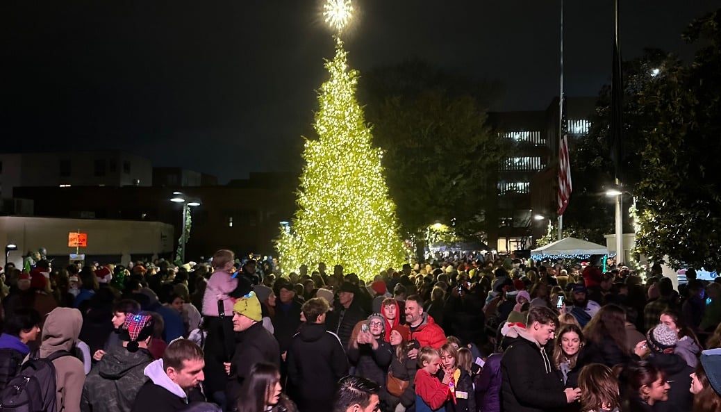 Crowd gathers around a brightly lit Christmas tree at a nighttime holiday event.