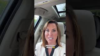 Woman in a white top and seat belt speaking from the front seat of a car.