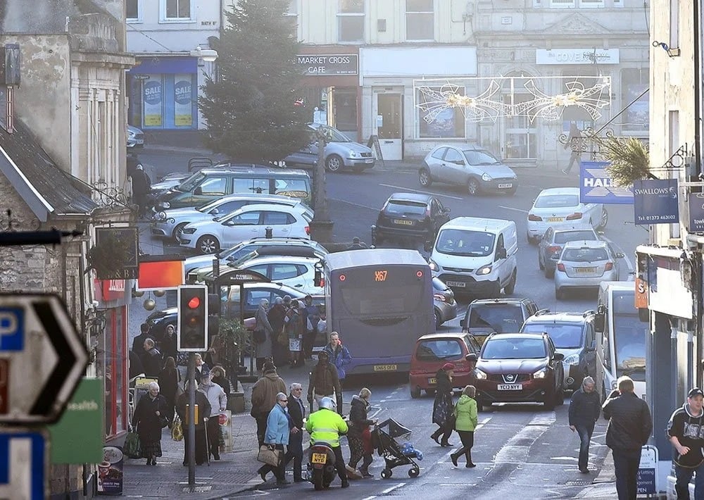 Busy town street with pedestrians, cars, and a bus amid shopfronts.