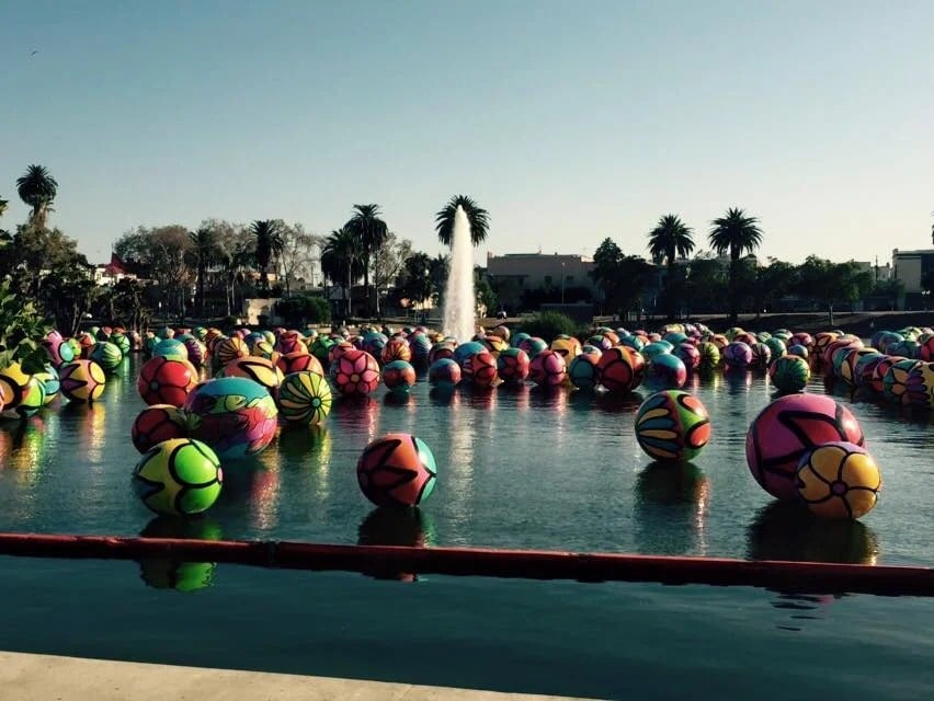 Colorful patterned spheres float in a park fountain pool, with palm trees and a central water jet behind them.