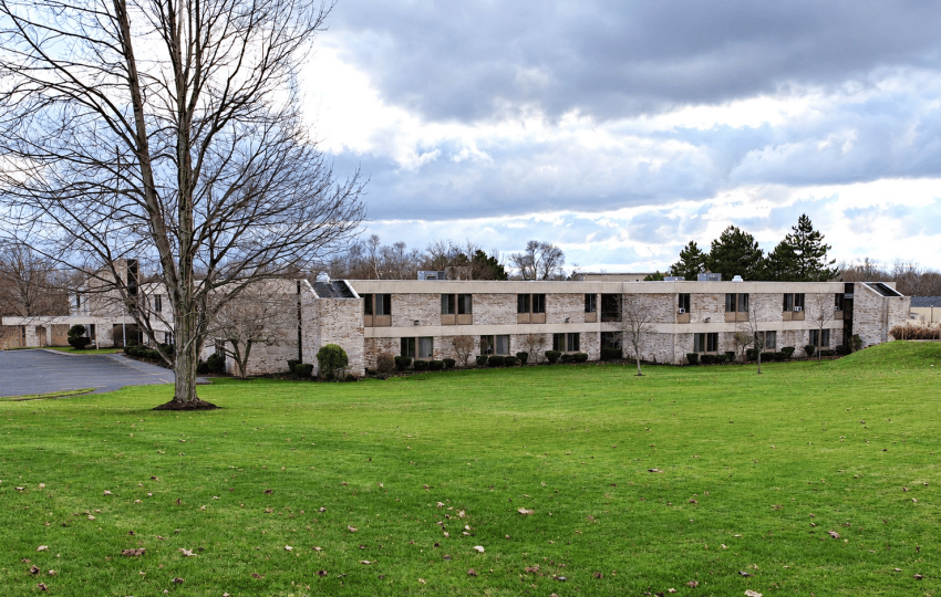 Low-rise senior living building beyond a broad lawn, with a leafless tree in front.